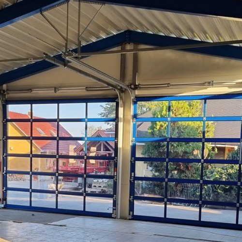 Két darab nagyméretű, teljesen üvegezett panoráma ipari garázskapu kék színű vázzal és sínrendszerrel egy modern autószerelő műhelyben vagy csarnokban – Koffenstein Kaputechnika Kft.